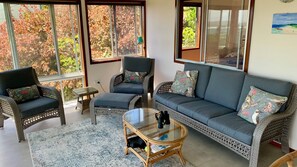 A cozy living room with wicker furniture, a glass coffee table, and large windows offering a view of trees.