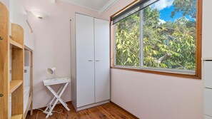 A room with a wardrobe, a desk with a lamp, and a window with a view of greenery.