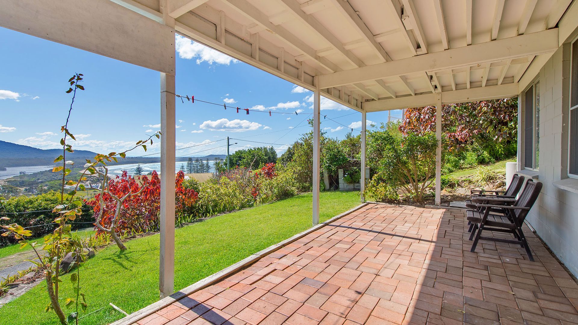 A covered patio with a view of the ocean and hills, featuring a brick floor and outdoor furniture.