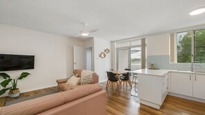 A modern living room with a sofa, a dining area with chairs, a kitchen with a countertop and cabinets, and a television mounted on the wall.