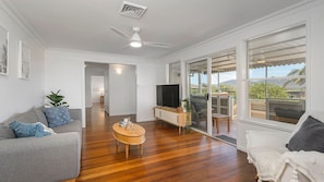 A spacious living area with a ceiling fan, a flat-screen TV on a wooden stand, a grey sofa, a round wooden coffee table, and a large sliding glass door leading to an outdoor patio.