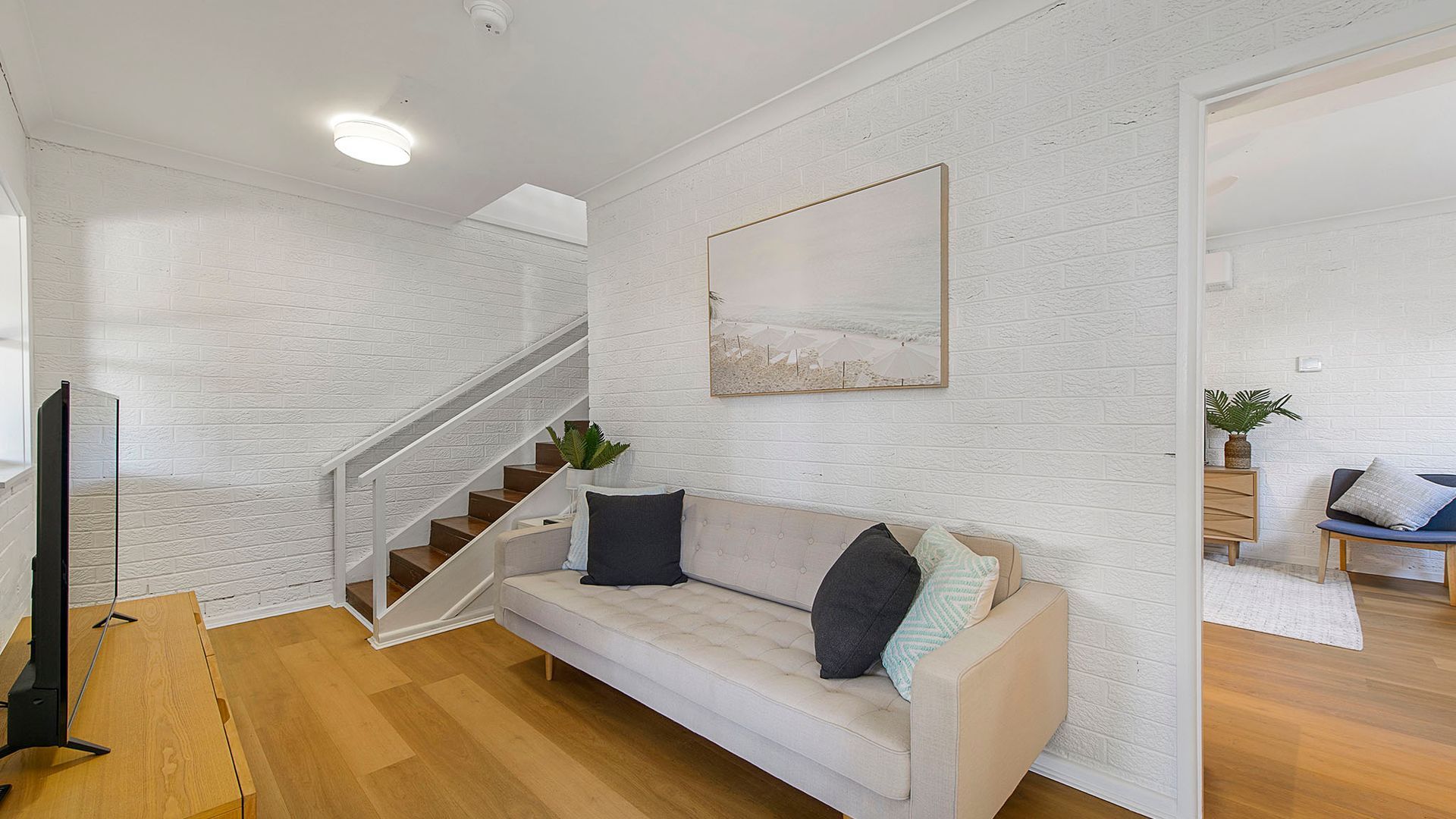 A living room with a white brick wall, a wooden staircase, a beige sofa with cushions, a flat-screen TV, and a framed picture.