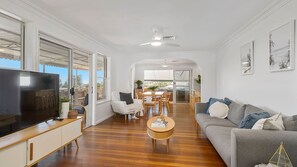 A modern living room with a flat-screen TV, a grey sofa, wooden flooring, and a dining area with a table and chairs.