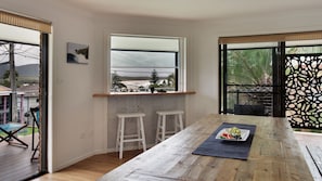 Uma área de refeições com mesa de madeira, dois bancos altos brancos, uma janela com vista para o exterior e um painel decorativo.