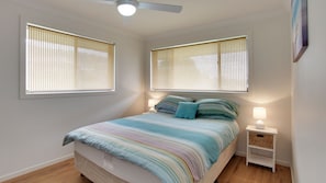 Quarto com cama, duas mesas de cabeceira, ventoinha no teto e janelas com cortinas.