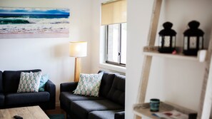 A living room with two sofas, a lamp, a painting of the ocean, and a window with blinds. - Seapoint Holiday House, 4 Gowing Street (Crescent Head)