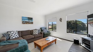 Una sala de estar con un sofá marrón, una mesa de centro de madera, un sillón verde y un ventanal con vistas a árboles y una casa.