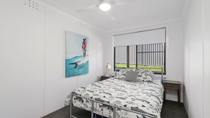 Un dormitorio con cama, mesita de noche, lámpara, un cuadro, ventana con persianas y ventilador de techo.