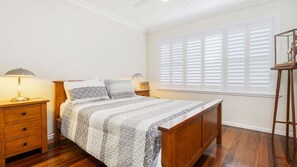A bedroom with a wooden bed, a nightstand with a lamp, a dresser, and a window with blinds. - Ondine, 4 Belmore Street (CRESCENT HEAD)