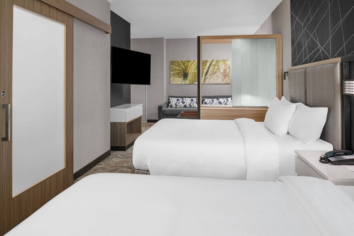 A hotel room with a large bed, a bedside table with a phone, a TV mounted on the wall, a sofa with patterned cushions, and a sliding door leading to a balcony.
