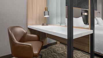 Quarto de hotel moderno com uma poltrona de couro, uma escrivaninha branca, um espelho e uma cama com roupa de cama branca.