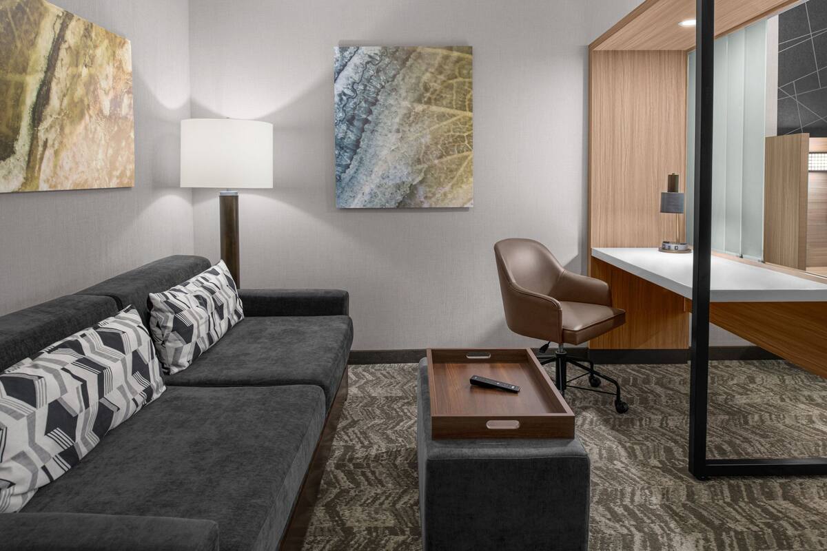 A modern hotel room with a grey sofa, a wooden side table with a remote control, a wooden desk, and a large mirror.