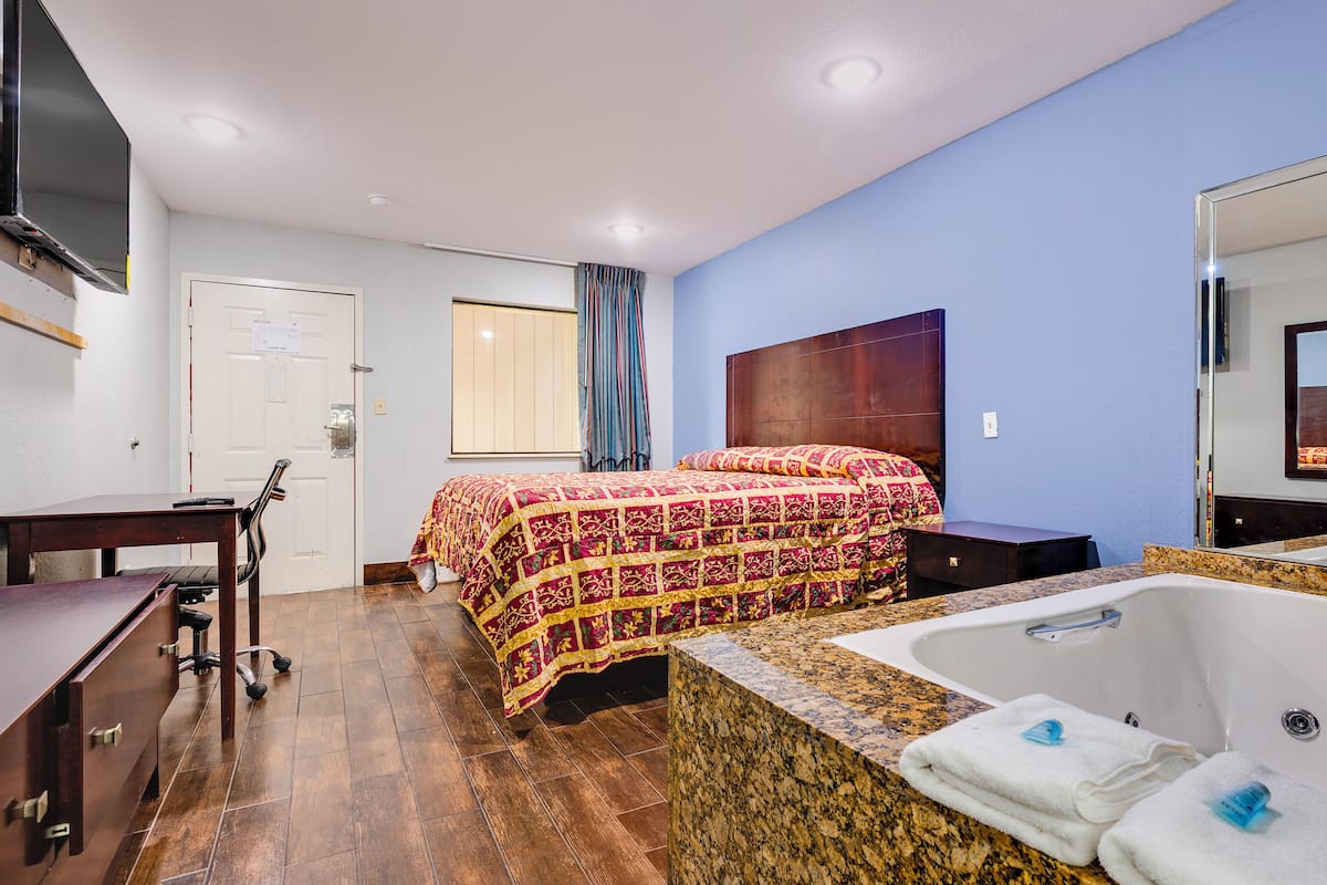 A hotel room with a bed, a desk, a chair, a large window with curtains, a bathroom with a bathtub, and a mirror.