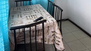 Een knus eetgedeelte met een tafel en stoelen, een raam met een plank vol decoratieve spullen, en een ventilatie rooster in het plafond.