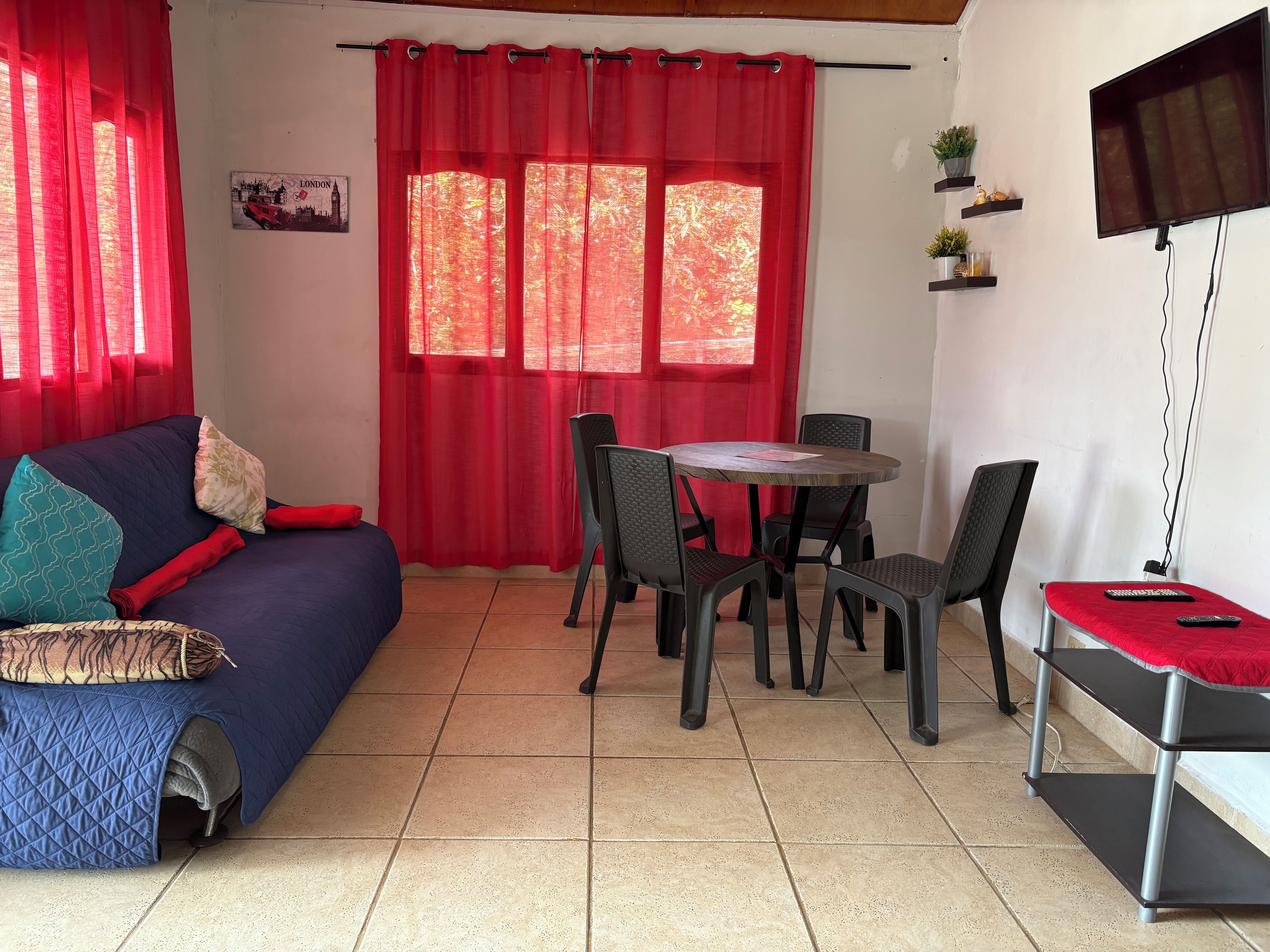 Une pièce avec un canapé bleu, des rideaux rouges, une table à manger avec des chaises, un téléviseur fixé au mur et une petite étagère avec des plantes.