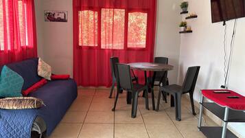Une pièce avec un canapé bleu, des rideaux rouges, une table à manger avec des chaises, un téléviseur fixé au mur et une petite étagère avec des plantes.