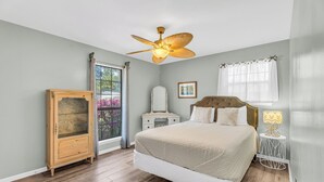 Une chambre à coucher comprenant un lit, un ventilateur de plafond, une commode, une table de chevet, une fenêtre avec des rideaux et un tableau encadré accroché au mur.