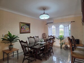 A dining area with a wooden table and chairs, a ceiling fan, a wall-mounted air conditioner, and a window with curtains.