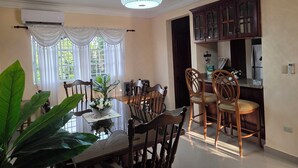 A dining area with a table, chairs, a ceiling fan, and a view of the outdoors through a window with curtains. - Secure Family-Oriented One of a Kind Condo (Cotui)