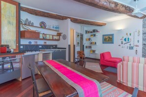 A modern kitchen with wooden beams, a dining area with a long table, and a living room with a red chair and striped sofa.