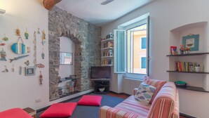 Un salón acogedor con pared de piedra, un sofá a rayas, cojines rojos, una estantería y un televisor.