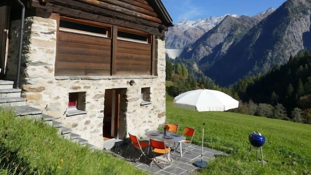 A stone cabin with a wooden roof, a patio with outdoor seating, and a mountain backdrop.