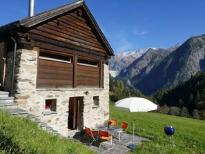 Een stenen chalet met een houten dak, een terras met buitenmeubels en een berg op de achtergrond.