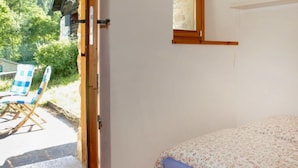 A bedroom with a bed, a window, and a door leading to an outdoor area with a chair and greenery.