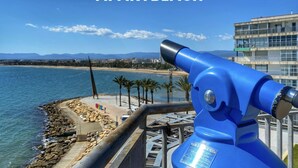Un telescopio azul con vista a una playa, montañas y un edificio al fondo.