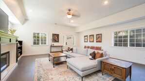 A spacious living room with a large sectional sofa, a coffee table, and a fireplace. There are multiple windows providing natural light and a ceiling fan. - Swanky Downtown Cowboy (Paso Robles)