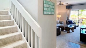 A staircase with a sign on the wall, a living room with a sofa, a coffee table, and a view of the outdoors. - Makanui 522 – Mountain Views & PBAC Access (Koloa)