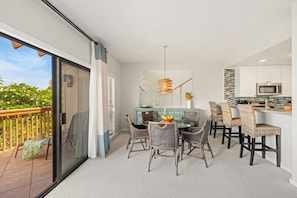 A dining area with a table and chairs, a kitchen with modern appliances, and a balcony with a view of greenery. - Makanui 522 – Mountain Views & PBAC Access (Koloa)