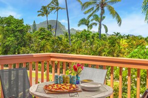 A wooden deck with a table set for a meal, featuring a pizza and canned drinks, overlooking a tropical landscape with palm trees and mountains.