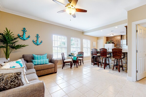 A spacious living room with a sofa, a dining table with chairs, and a kitchen in the background. - Coastal-themed home near the ocean with pool, AC & fast WiFi (South Padre Island)