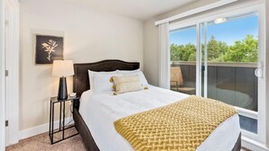 Une chambre avec un grand lit, une table de chevet avec une lampe, un tableau encadré au mur et un balcon donnant sur la verdure.