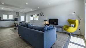 Un salon moderne avec un canapé, un fauteuil jaune, un téléviseur et, au fond, un coin cuisine.