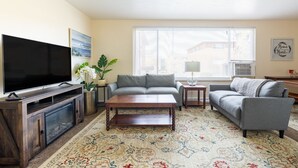 Ein Wohnzimmer mit Flachbildfernseher, grauen Sofas, einem Holztisch, einem gemusterten Teppich und einer Topfpflanze.