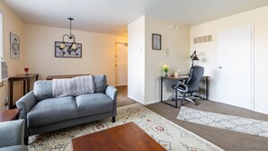 Ein Wohnzimmer mit grauem Sofa, einem Holztisch und einem gemusterten Teppich. Es gibt einen Schreibtisch mit Stuhl und Lampe.