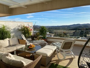 A balcony with a sofa, chairs, a table with fruit, and a view of the countryside.