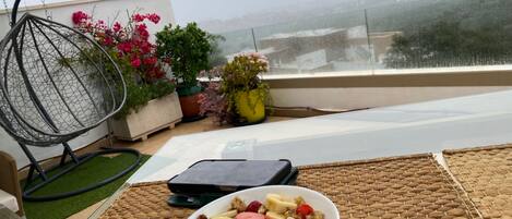 Un comedor con una mesa lista para el desayuno, que incluye un tazón de cereal con frutas, una silla y plantas en macetas.