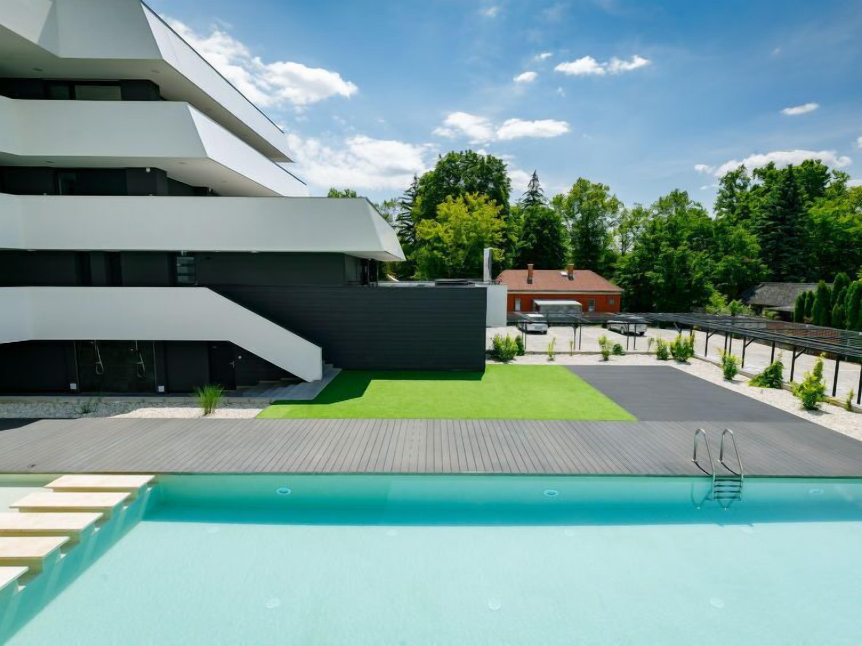 Un edificio moderno con zona de piscina, que incluye una terraza de madera y una piscina azul cristalina.