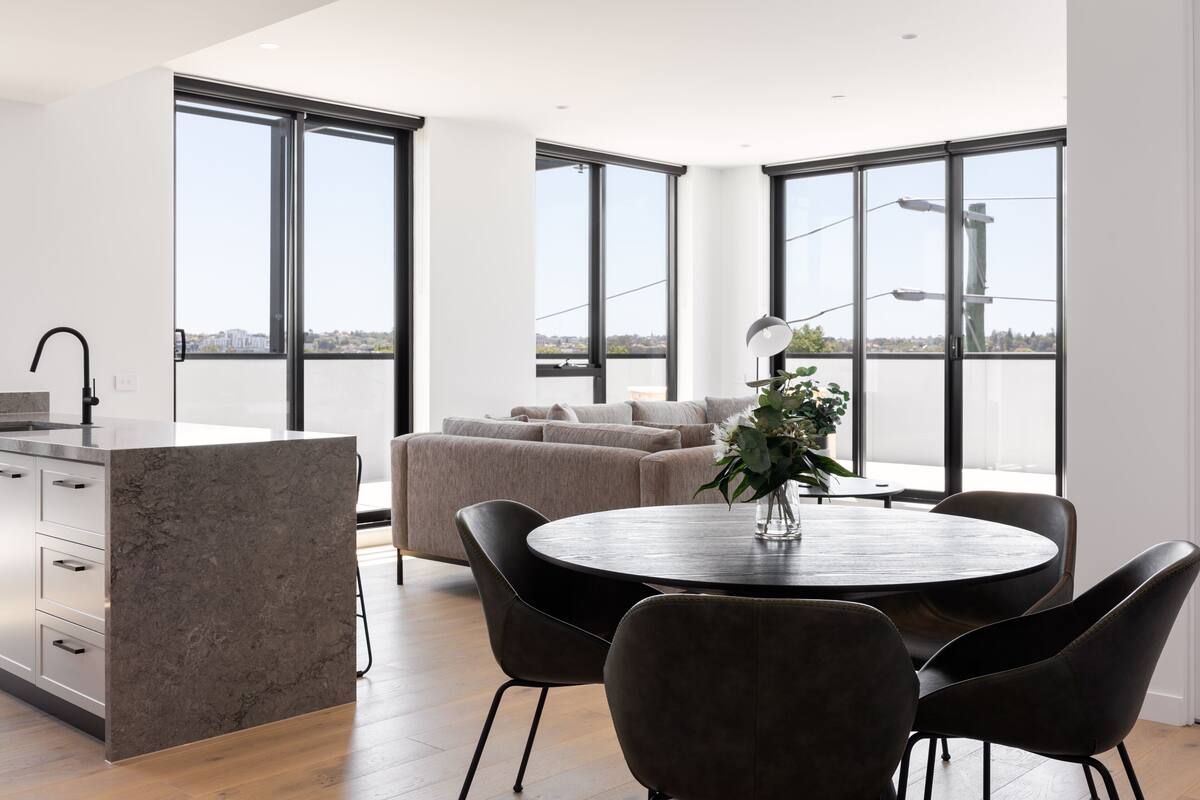 A modern kitchen with a marble island, a dining area with a round table and chairs, and a large window offering a city view.