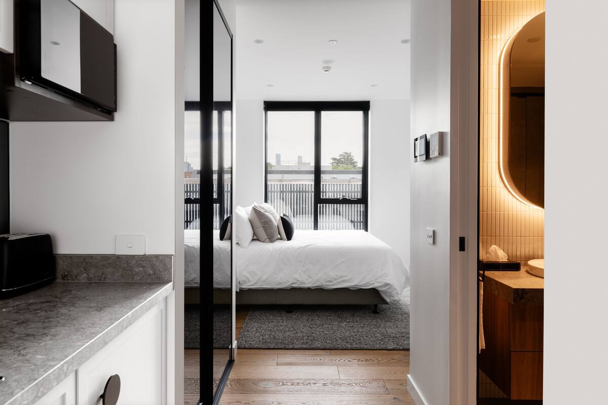 A modern bedroom with a large window, a bed with pillows, a bedside table, and a view of the cityscape.