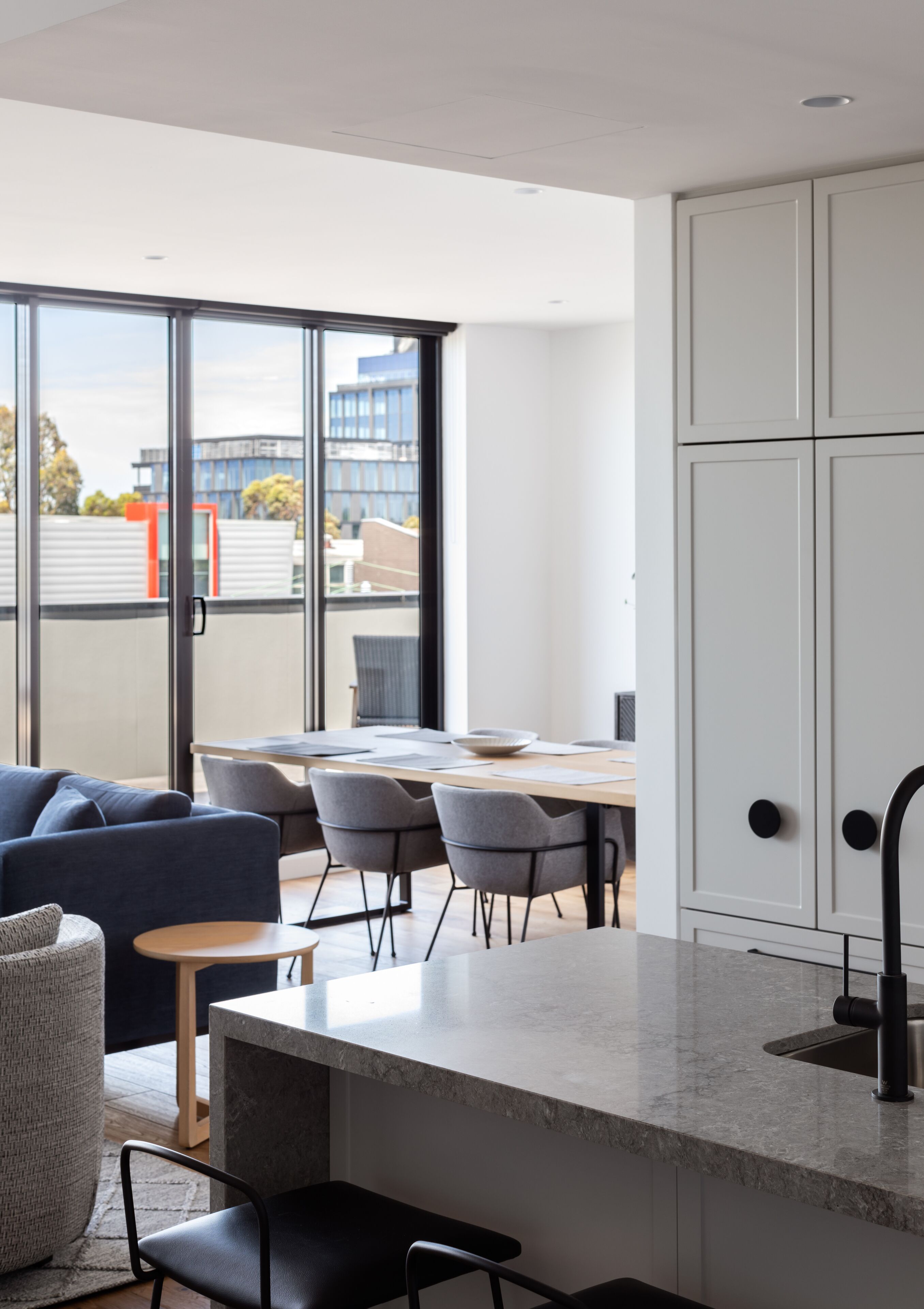 Cozinha moderna com bancada de mármore, pia integrada e armários brancos. Há uma área de jantar com mesa e cadeiras, e um sofá. Janelas amplas oferecem vista para a cidade.