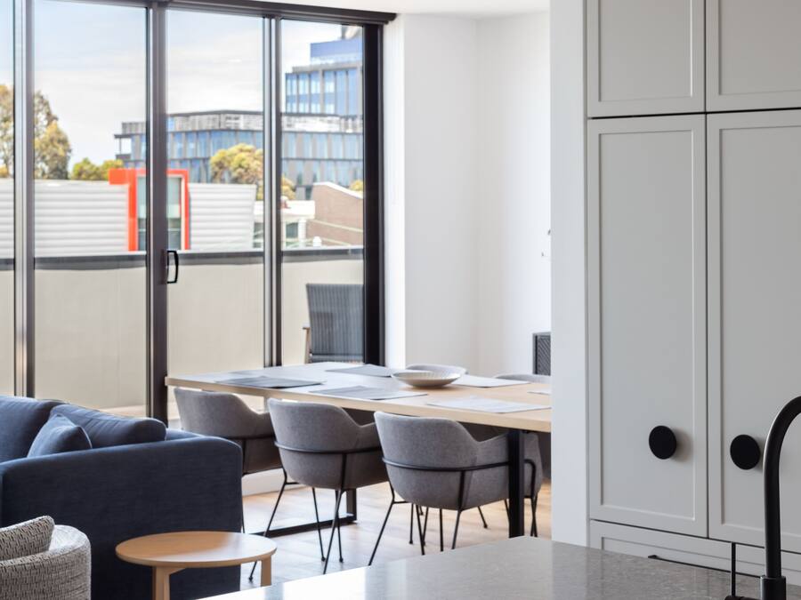 Cocina moderna con encimera de mármol, lavamanos integrado y armarios blancos. Hay un comedor con mesa y sillas, y un sofá. Amplios ventanales ofrecen vistas de la ciudad.