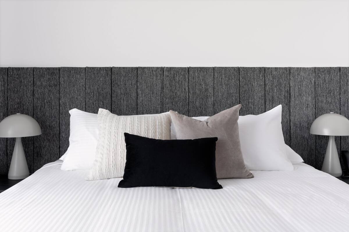 A neatly made bed with a white bedspread and multiple pillows, including a black one, against a wooden headboard.