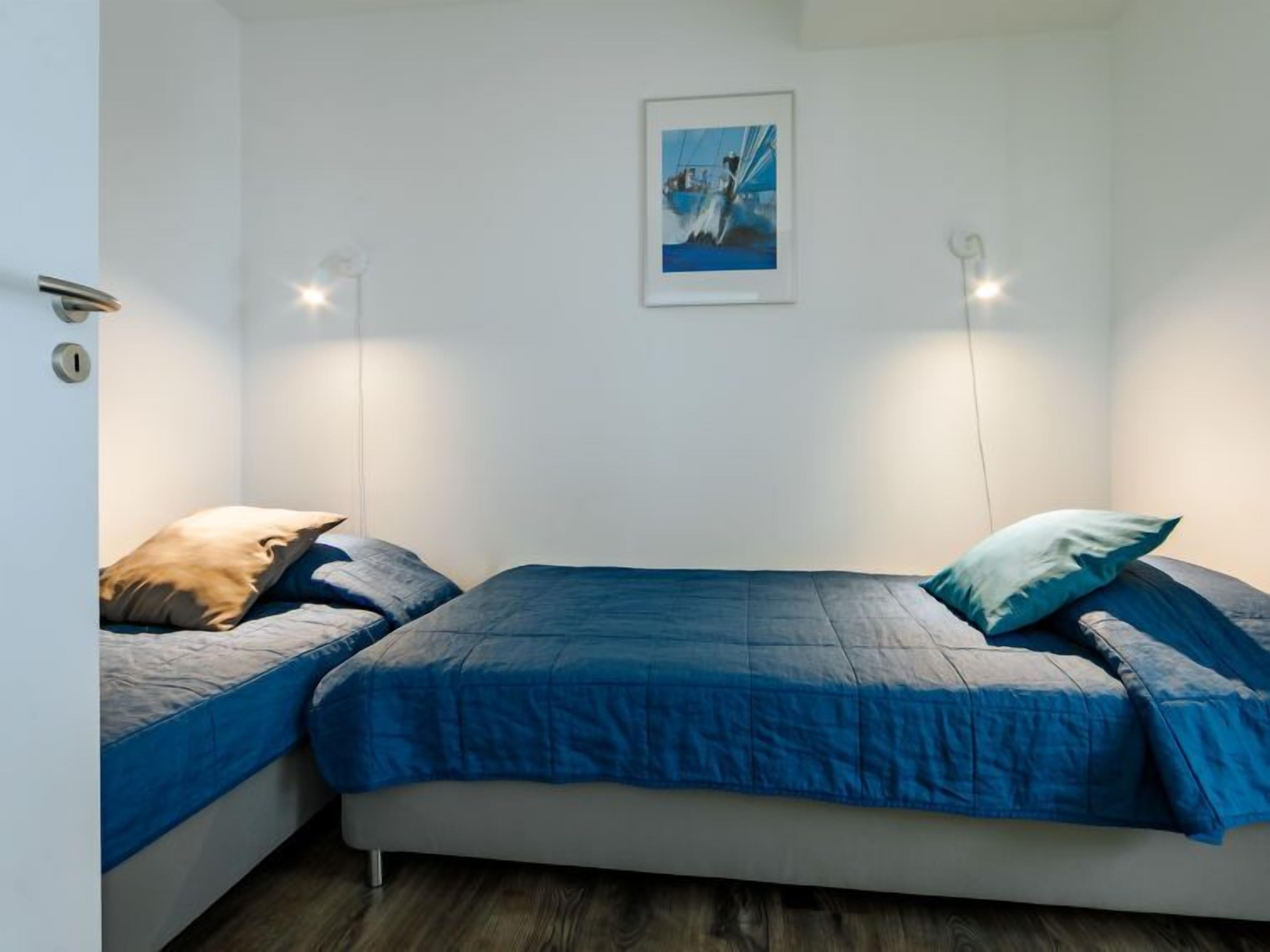 A bedroom with two beds, each with blue bedding. There is a framed picture on the wall and two wall-mounted lights.