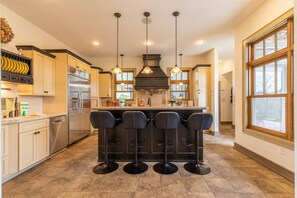 A modern kitchen with stainless steel appliances, a central island with bar stools, pendant lighting, and large windows.