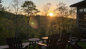 An outdoor seating area with chairs and tables, overlooking a forested landscape with a building in the background.