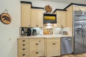 A kitchen with beige cabinets, a stainless steel refrigerator, a coffee maker, a toaster, and a wall-mounted shelf with decorative items.
